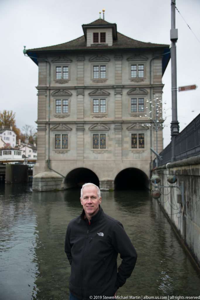 Man centered before Zürich Rathaus, with its symmetrical stone façade and river arches visible across the Limmat.