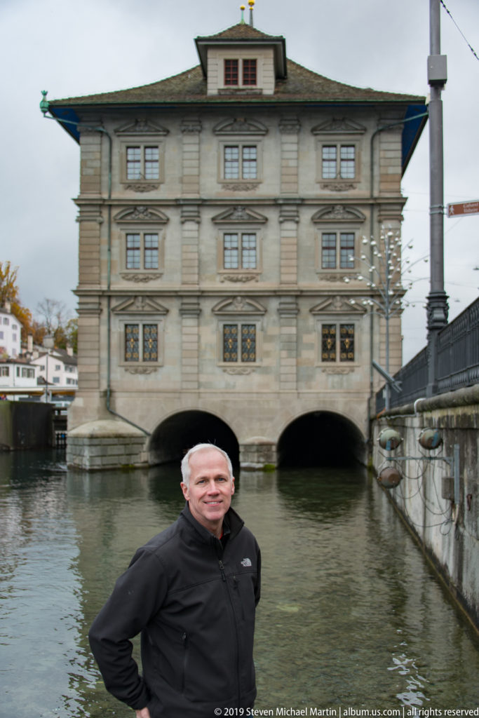 Man standing beside the Limmat with Zürich Rathaus, a pale stone building on broad river arches, behind him.