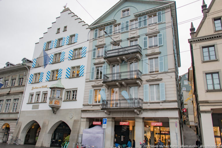 Adjacent Zurich historic façades with striped shutters, stepped gable, pale blue shutters, balconies, arcades, and shopfronts.