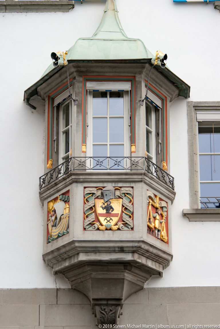 Projecting Zurich oriel window with green copper roof, iron rail, red-and-gold trim, and painted heraldic panels.