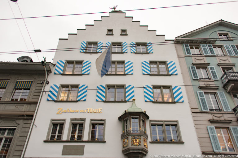 White stepped-gable Zunfthaus zur Haue façade with blue-and-white shutters, flag, and projecting decorated oriel.