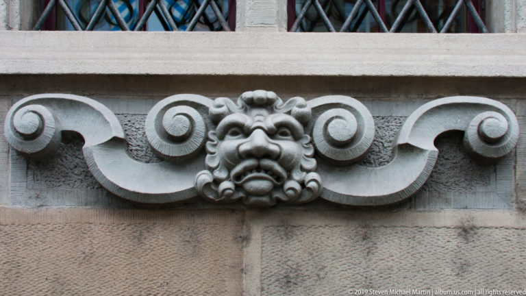 Carved stone grotesque face centered between symmetrical scrolling volutes on a Zurich building façade.