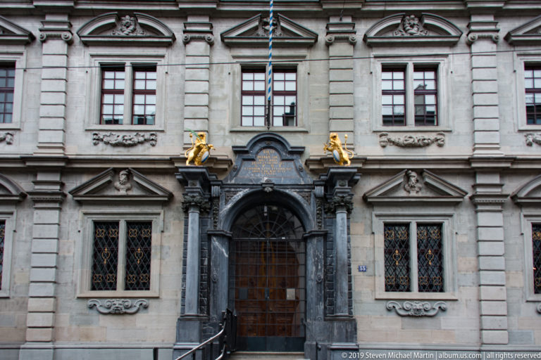 Ornate Zurich stone façade with dark arched doorway, carved heads, symmetrical windows, and two gilded lions above.