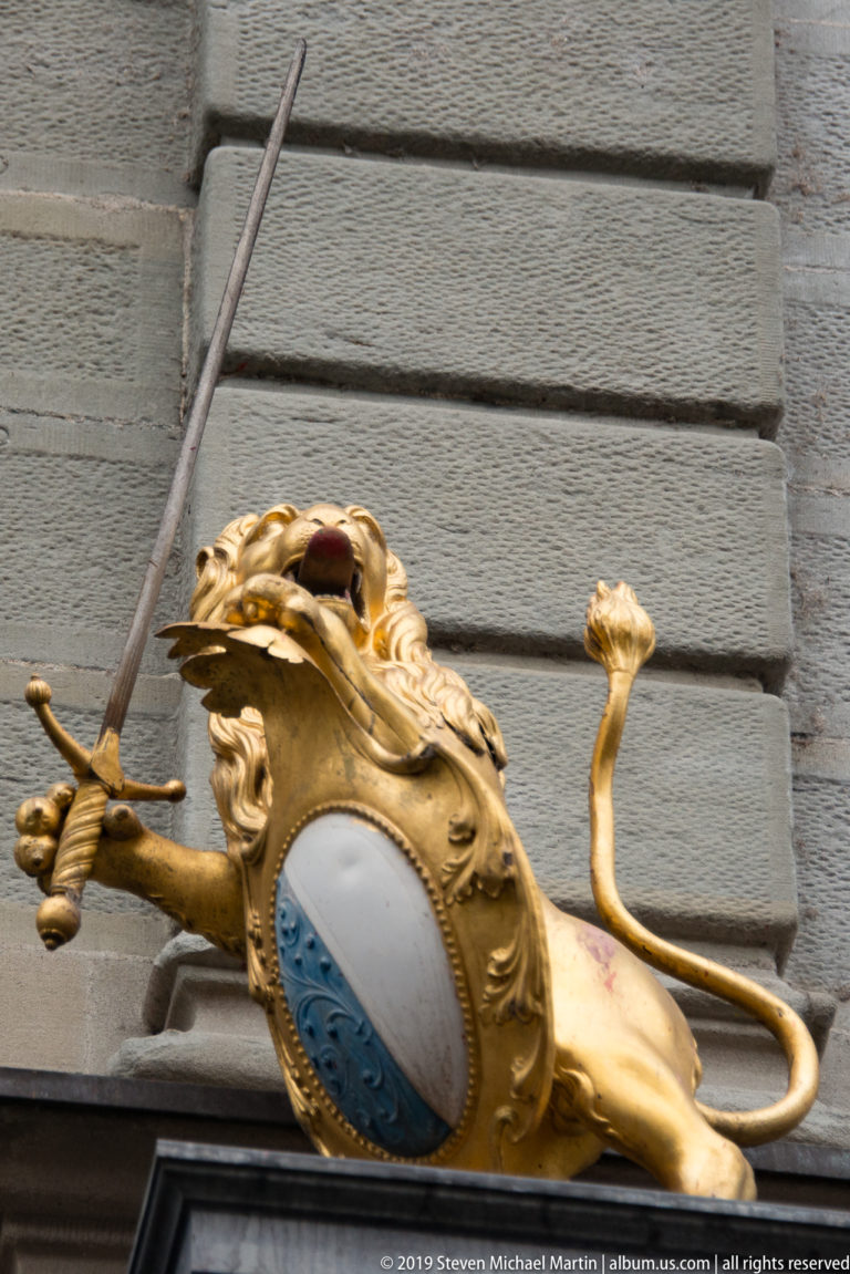 Gilded lion-shaped projecting sign holding an oval blue-and-white shield against a gray rusticated Zurich façade.