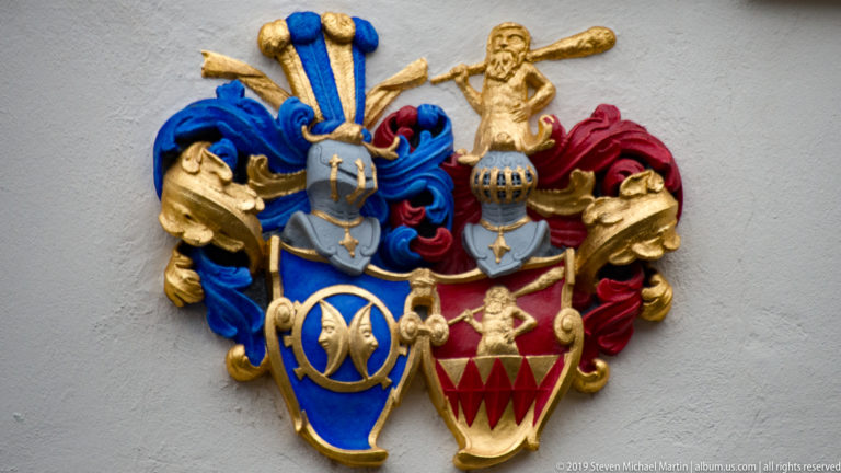 Paired blue and red heraldic shields with gold ornament, colorful mantling, and a small sculpted figure above.