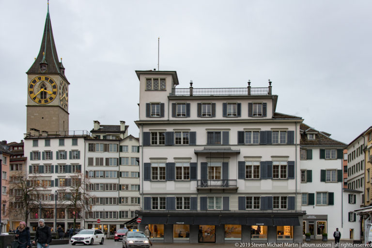 St. Peter’s large clock tower rising behind pale shuttered buildings, cars, and pedestrians in central Zurich.