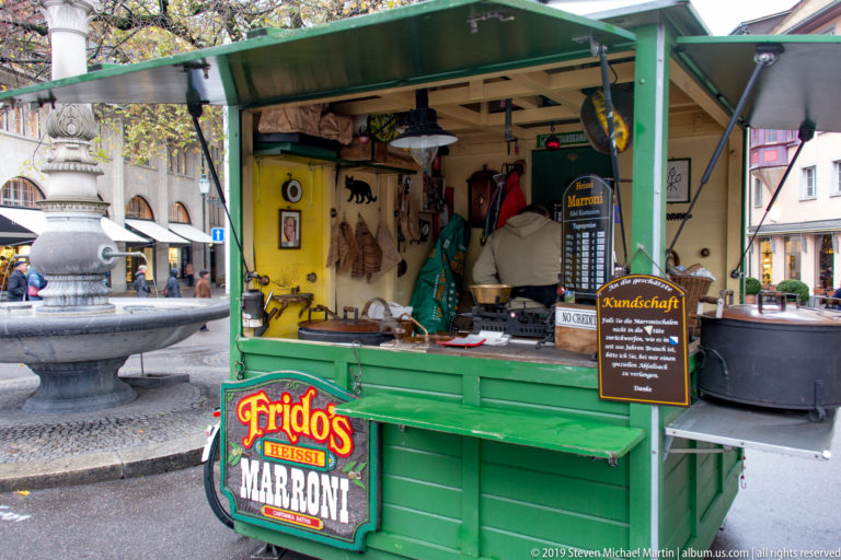 Green Zurich street stall advertising hot Marroni chestnuts, with menu boards, roasting equipment, and a stone fountain behind.