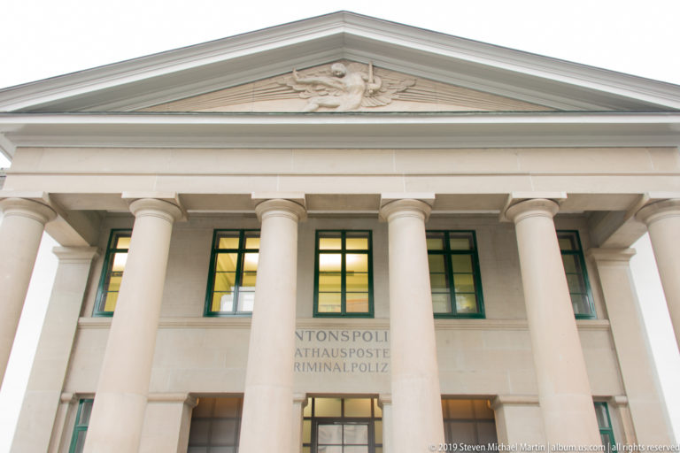 Neoclassical Rathauspost façade with four large columns, triangular pediment relief, pale stone, and green-framed windows.