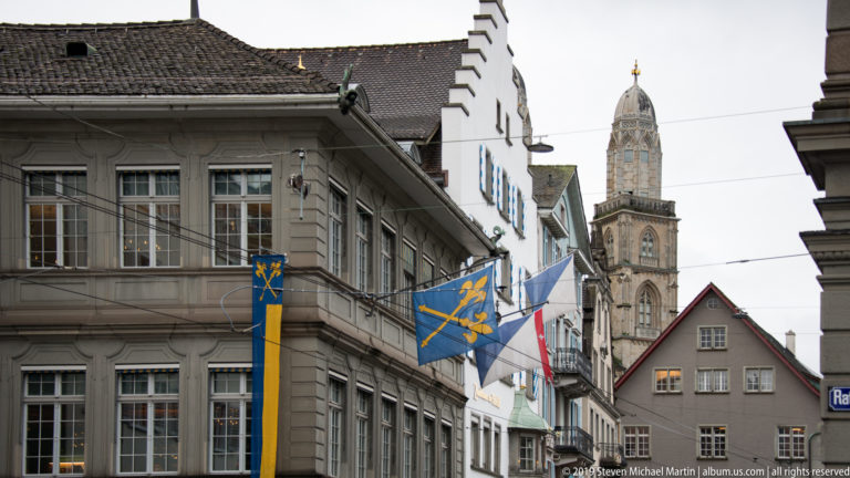 Heraldic flags on a gray Zurich building with a tall stone church tower rising beyond narrow Old Town façades.