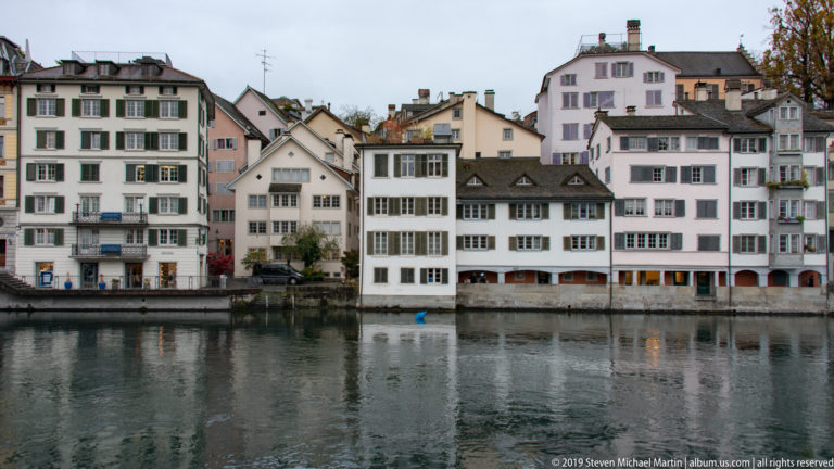 White and pastel historic houses with shuttered windows lining the opposite bank of the Limmat in Zurich.