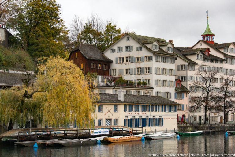 Historic Zurich riverfront houses with shutters, moored boats, a willow tree, and a slender green-roofed tower beside the Limmat.