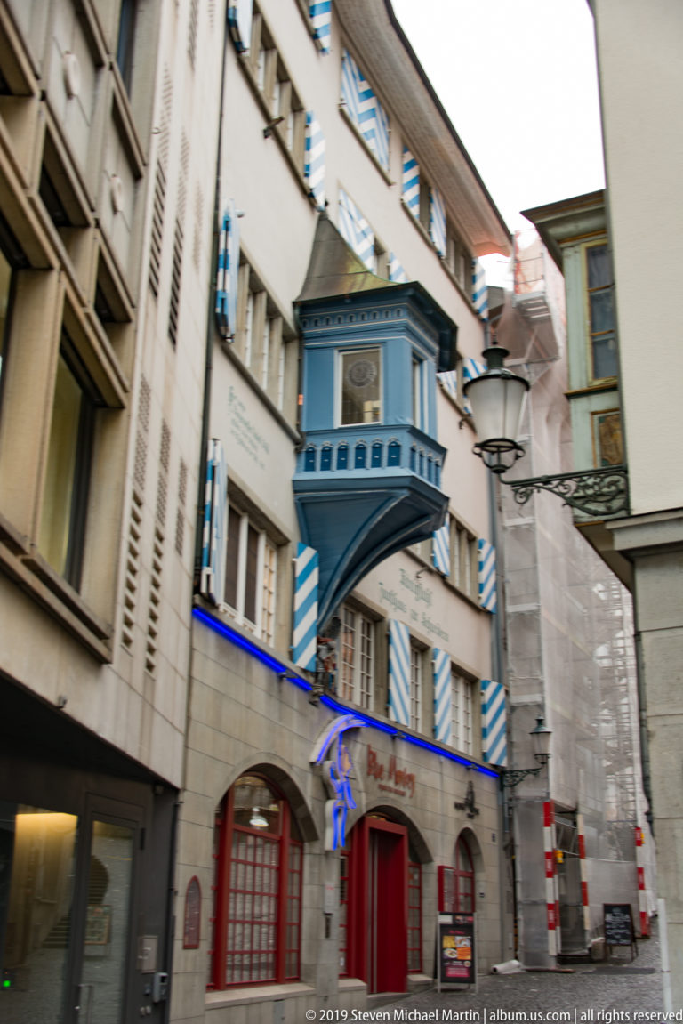 Blue projecting oriel, blue-and-white striped shutters, and red entrance on an ornate Zurich Old Town façade.