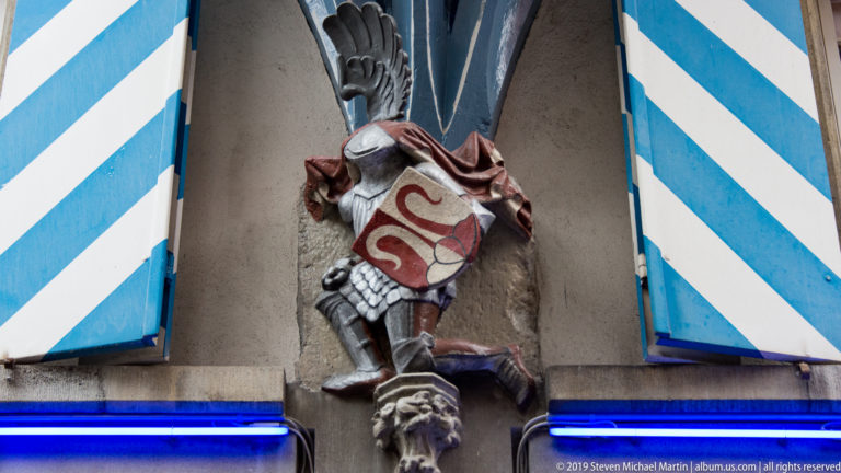Armored heraldic figure with painted shield mounted between blue-and-white striped shutters on a Zurich façade.