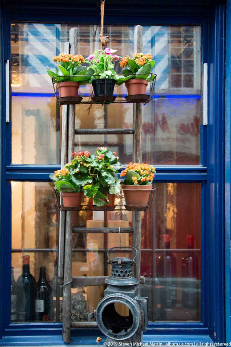 Tiered pots of colorful flowers arranged on a wooden stand outside a blue-painted storefront in Zurich.