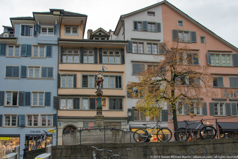 Painted figure fountain before pastel Zurich Old Town houses, with shuttered windows, bicycles, and autumn foliage.