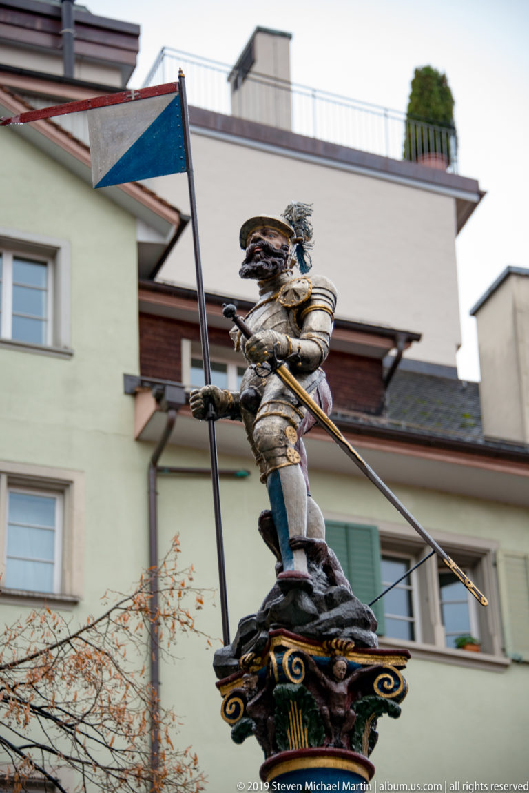 Painted armored standard-bearer holding a long pole atop an ornate fountain column, with modern Zurich buildings behind.