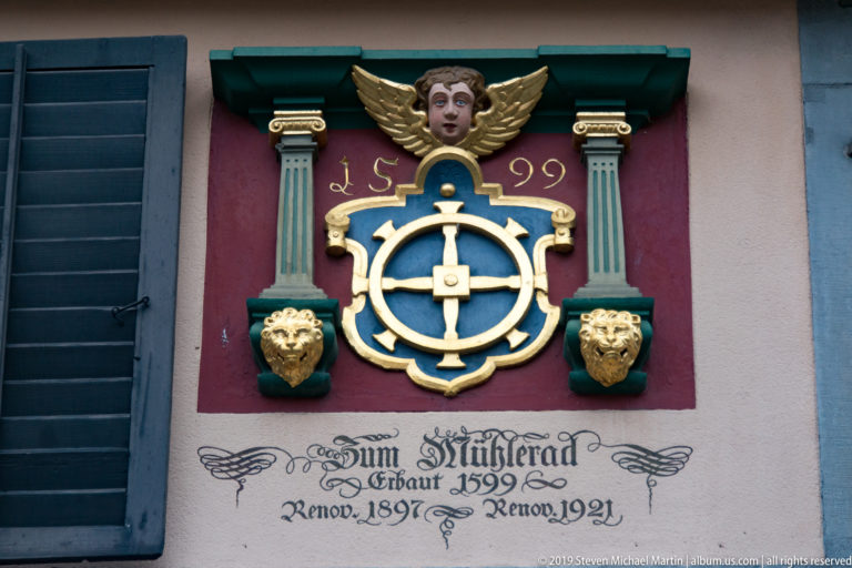 Gilded mill-wheel emblem on a red panel with columns, lion heads, winged head, and painted Zum Mühlrad renovation inscription.