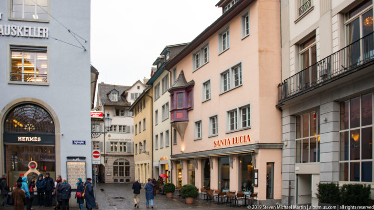 Pedestrian Zurich street with Santa Lucia and Hermès storefronts, pastel façades, a purple bay window, and people walking.