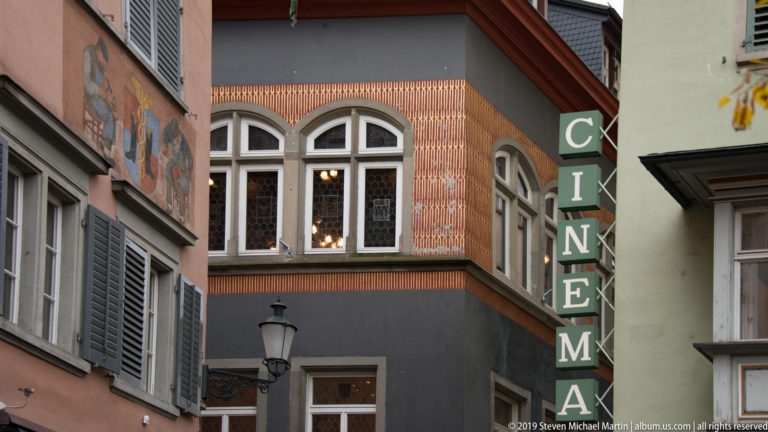 Vertical CINEMA sign between narrow Zurich façades with arched windows, patterned wall decoration, shutters, mural fragment, and lantern.
