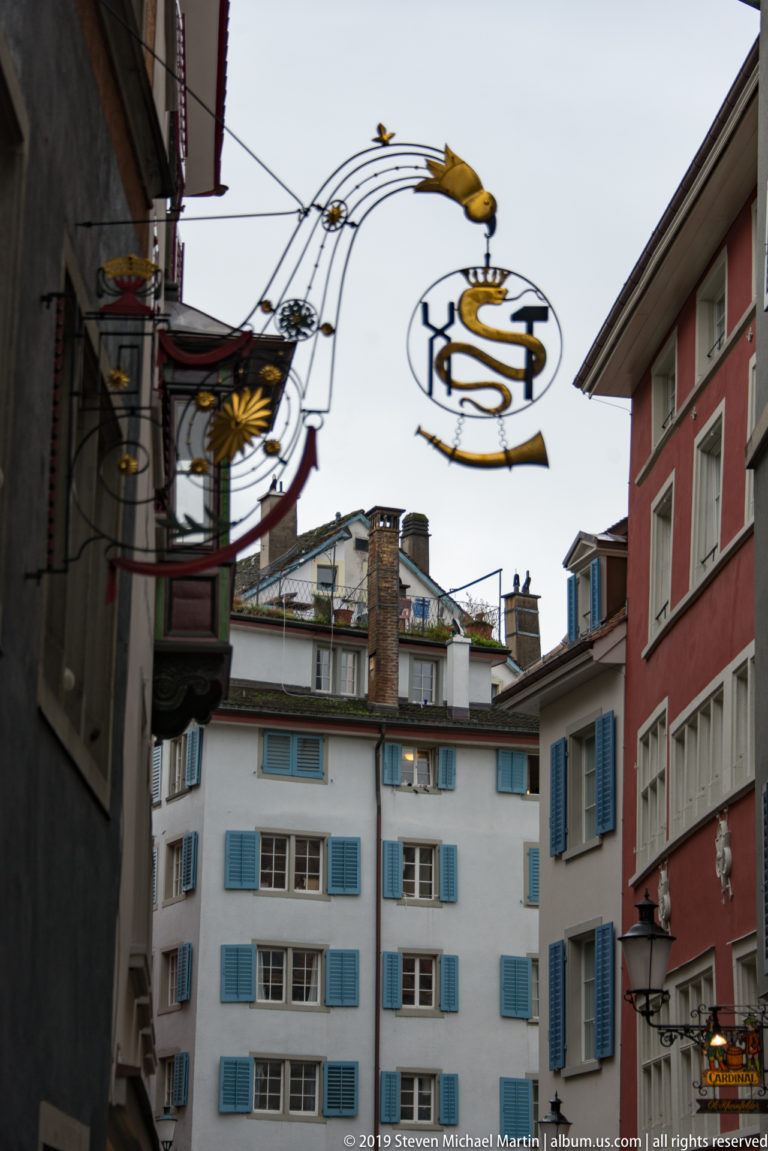 Gilded wrought-iron hanging signs suspended above a narrow Zurich street with pastel façades and blue shutters.