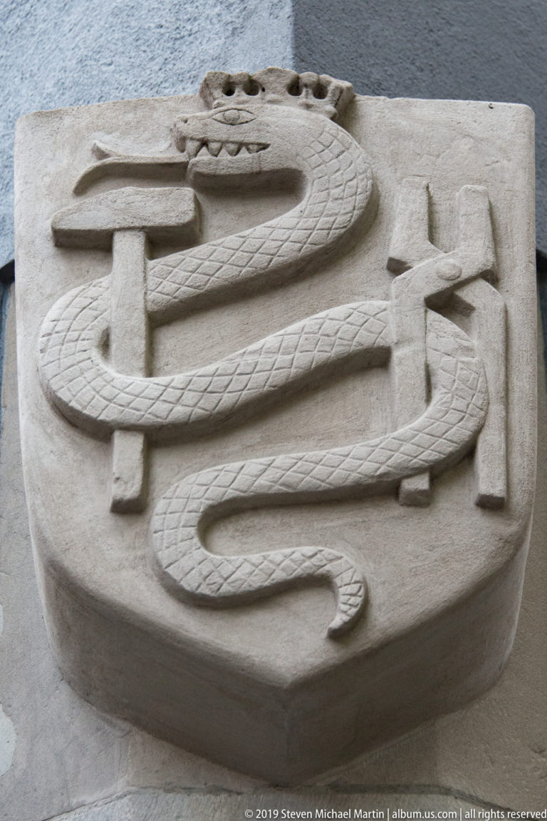 Pale stone relief of a long coiled serpent with incised scales looping across a shield-like panel on a dark wall.