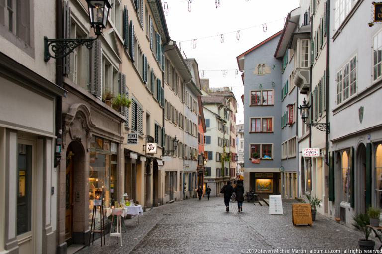 Cobblestone pedestrian lane in Zurich lined with shuttered historic buildings, shopfronts, projecting bays, signs, and several walkers.