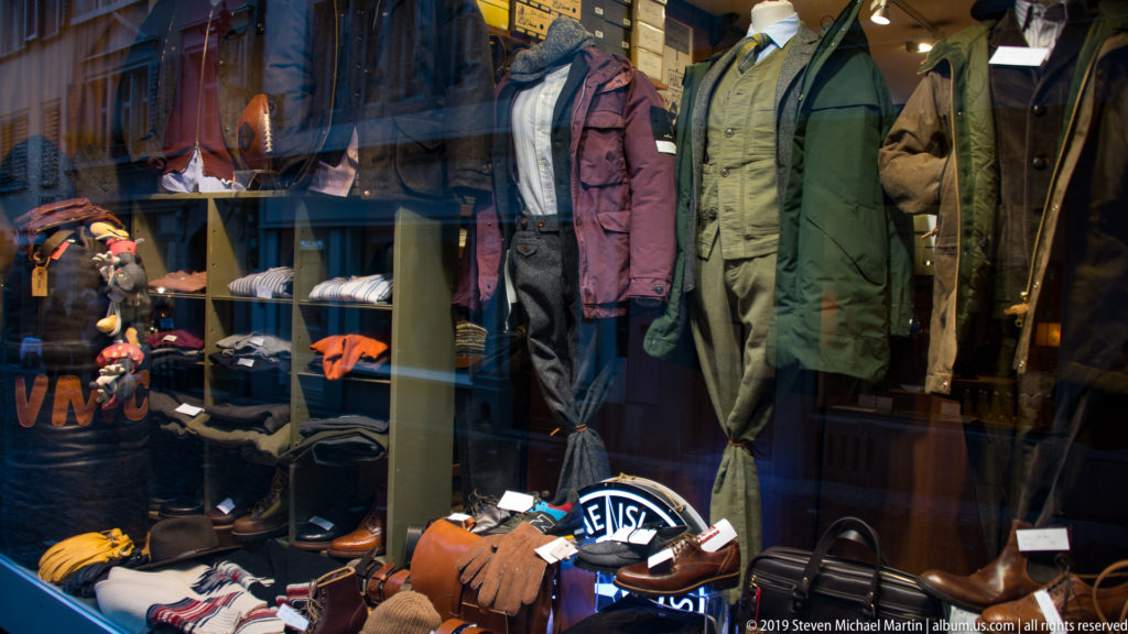 Menswear shop window with three mannequins in jackets above folded clothing, shoes, bags, and accessories behind reflective glass.