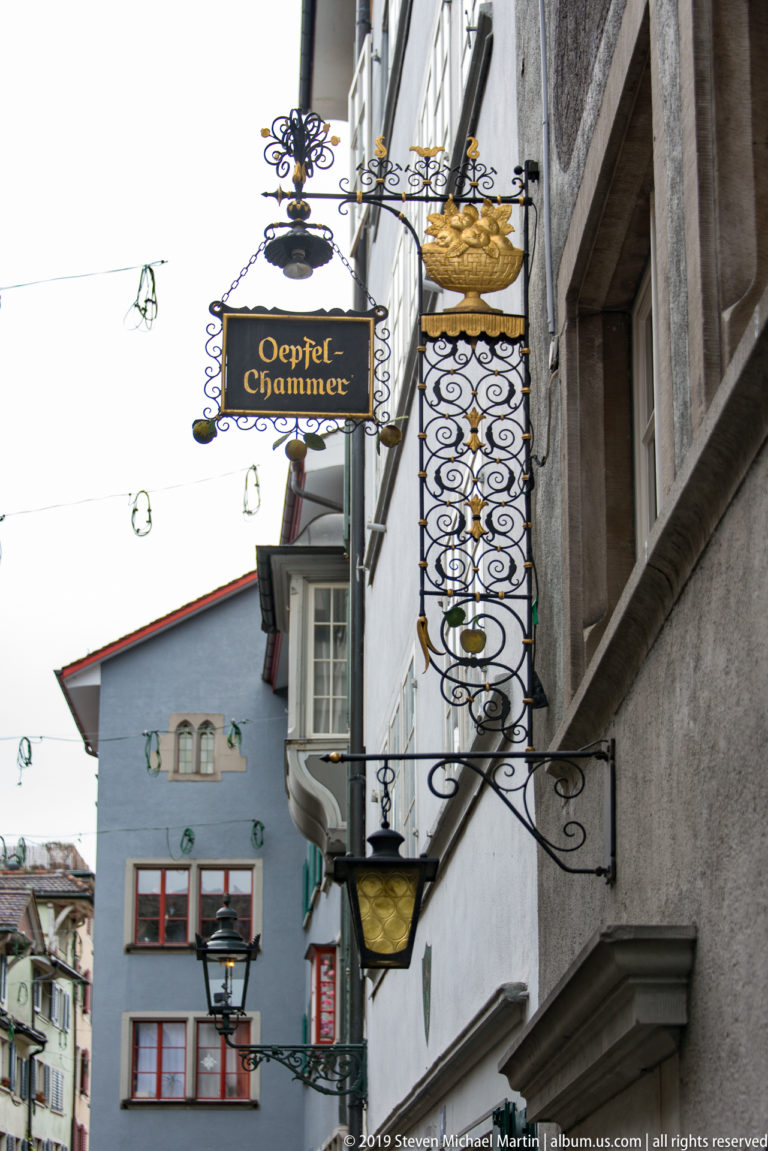 Wrought-iron Oepfel-Chammer sign and lanterns projecting above a narrow Zurich street lined with historic façades.