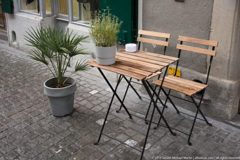 Two wooden folding chairs and a small table beside potted plants on a cobblestone lane against a gray wall.