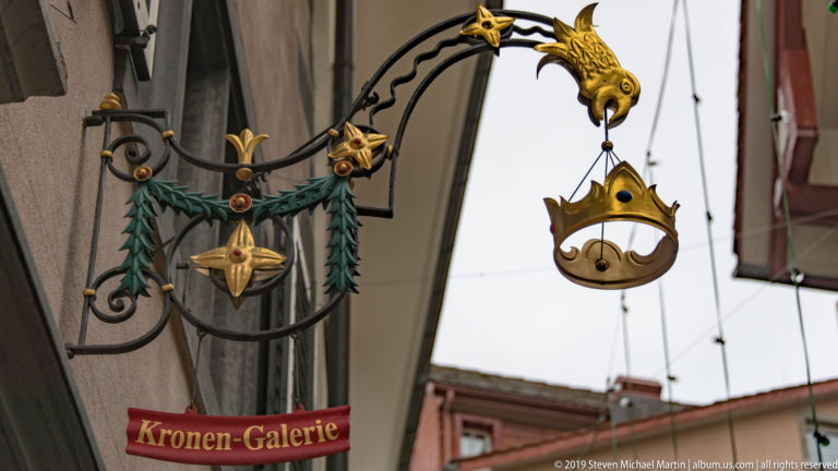 Ornate Kronen-Galerie hanging sign with gilded crown, dragon-like bracket, black iron scrollwork, and red name plaque.