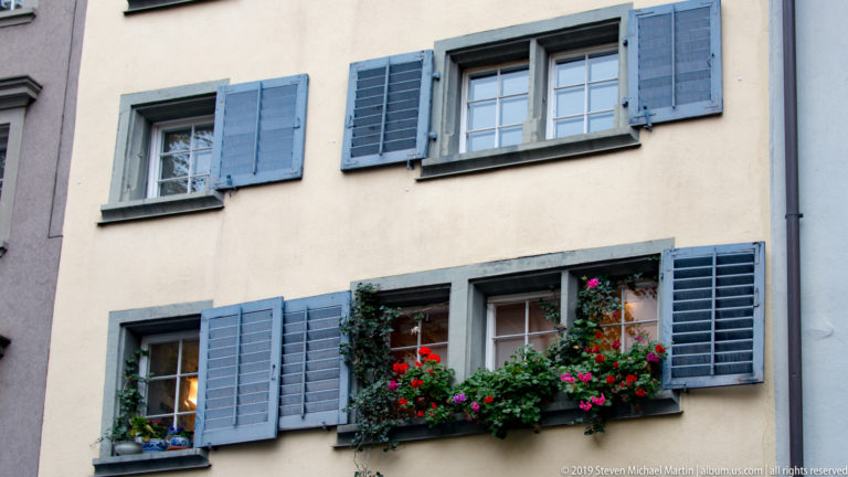 Pale Zurich façade with blue-gray shutters and divided windows, including a lower window box overflowing with red and pink flowers.