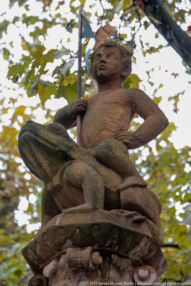 Weathered seated stone figure holding a long staff atop a fountain, framed by autumn leaves.
