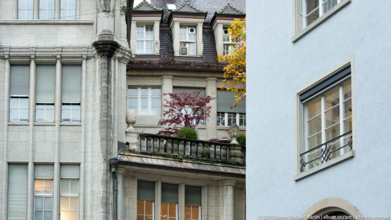 Stone terrace with balustrade, planters, and tall windows squeezed between an ornate gray façade and plain blue wall.