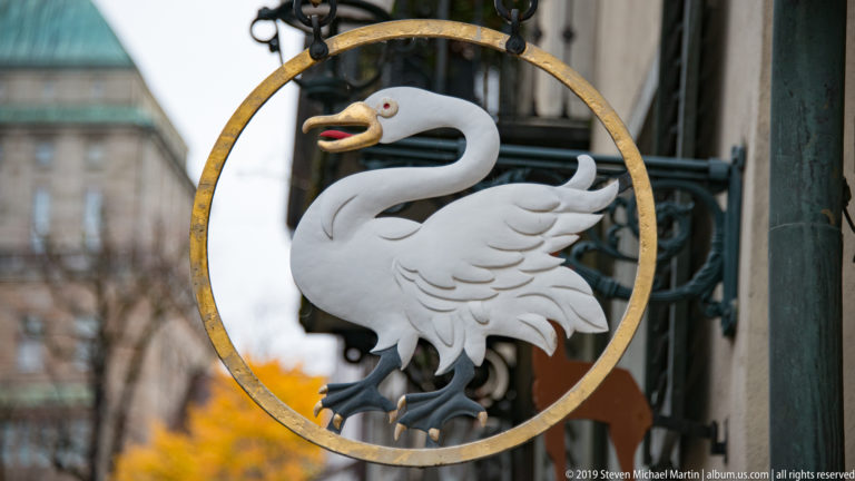 White swan with red beak suspended inside a gilded circular hanging sign against a softly blurred Zurich street.