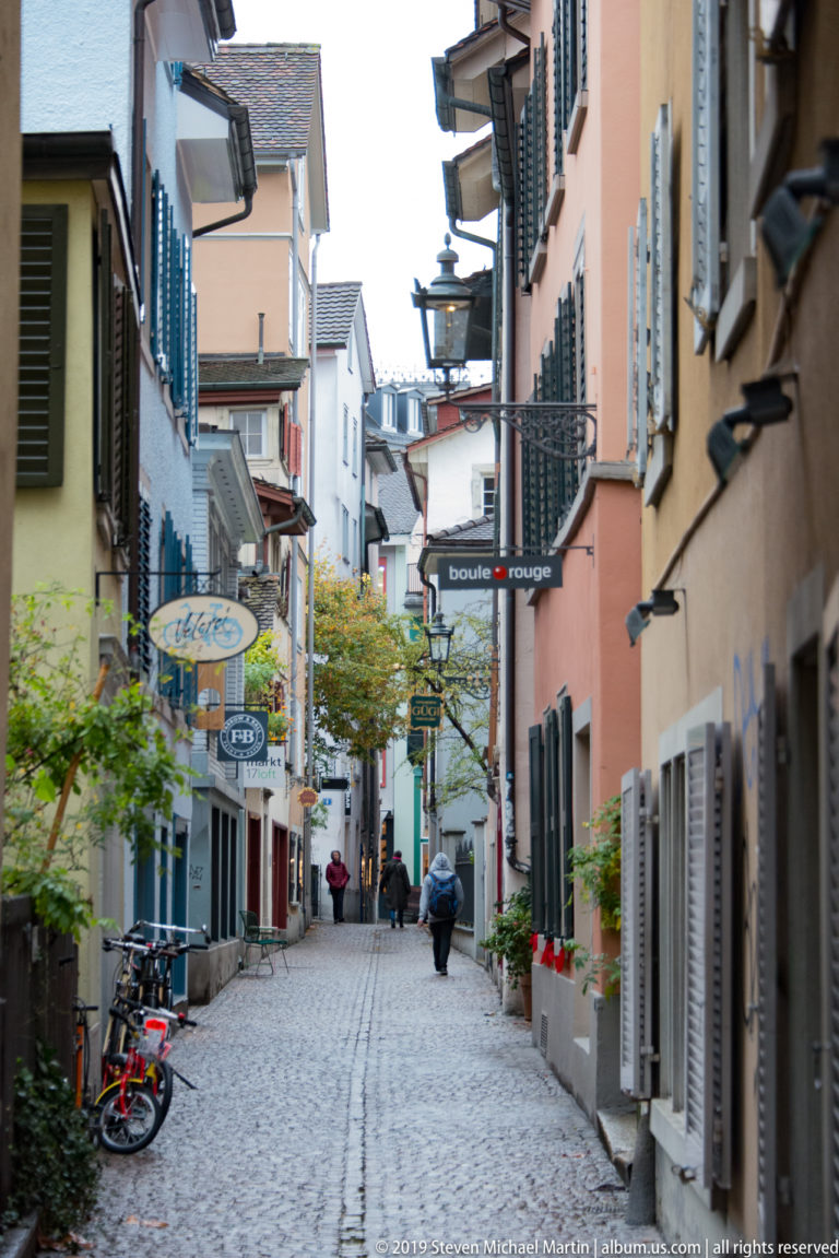 Narrow cobblestone Zurich lane lined with pastel buildings, shutters, shop signs, potted plants, bicycles, and pedestrians.