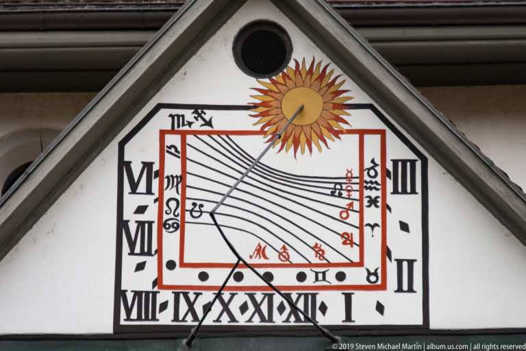 Triangular gable containing a painted sundial with Roman numerals, black hour lines, red markings, and a golden sun.