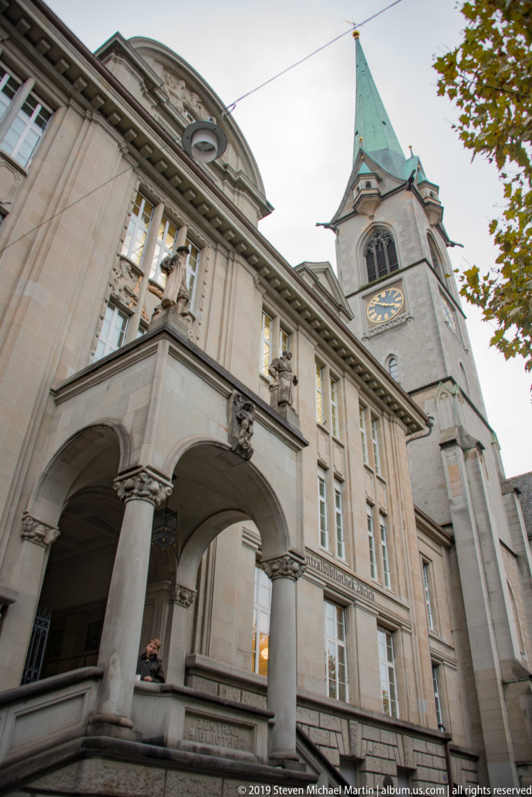 Stone civic building with columned loggia in the foreground and a clock-towered church with green copper spire rising behind.