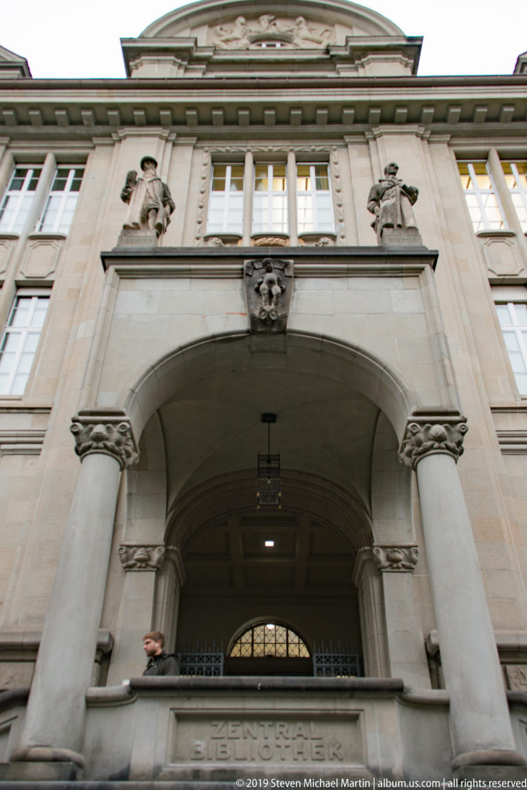 Low-angle view of a Zurich stone façade with deep columned loggia, carved heads, relief ornament, and partial Gesellschaft inscription.