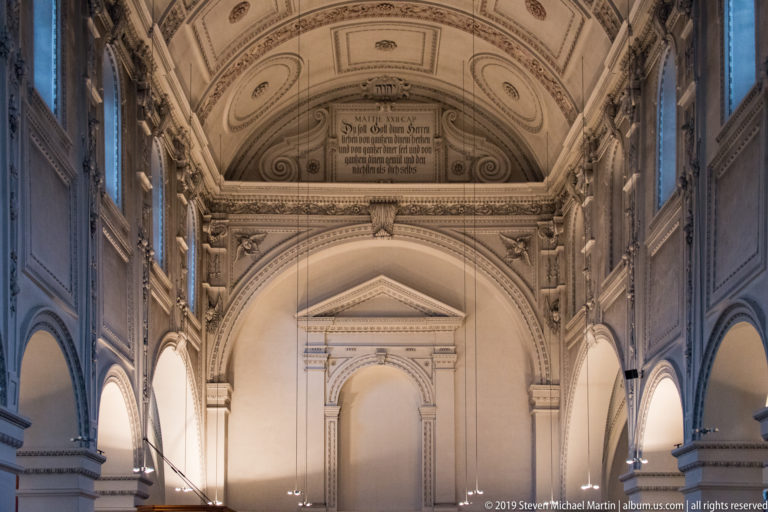 Pale monumental interior with side arcades, pilasters, coffered barrel vault, carved ornament, and a German inscription above the far arch.