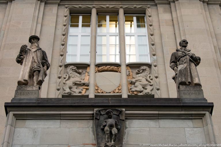 Stone statues labeled Gessner and Bodmer flanking an ornate shield and carved lions beneath tall windows on a Zurich façade.