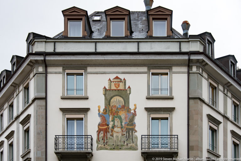 Pale Zurich building with symmetrical windows and balconies framing a colorful heraldic mural of figures beneath a fortified emblem.