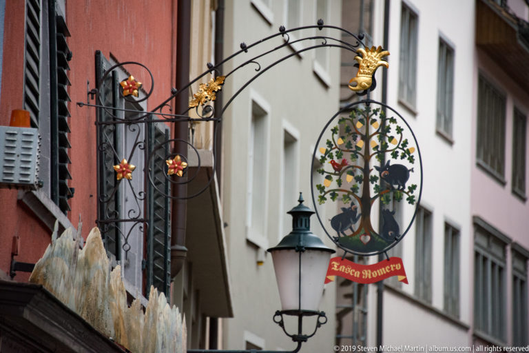 Ornate wrought-iron Andrea Romero hanging sign with gilded flowers, animal figures, painted tree, and lantern on a narrow street.