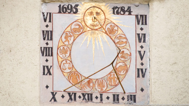 Painted wall sundial with Roman numerals, zodiac symbols, sun face, and the dates 1695 and 1784.