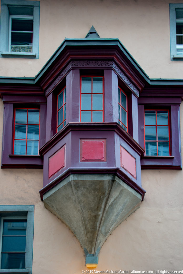 Purple polygonal oriel with red-framed windows and turquoise trim projecting from a pale Zurich façade.
