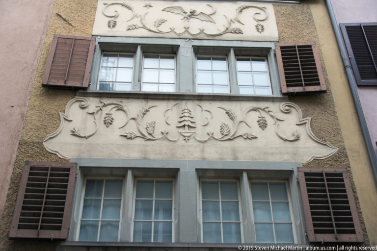 Shuttered windows on a Zurich façade separated by cream stucco reliefs of foliage, fruit, scrollwork, and a small tree.