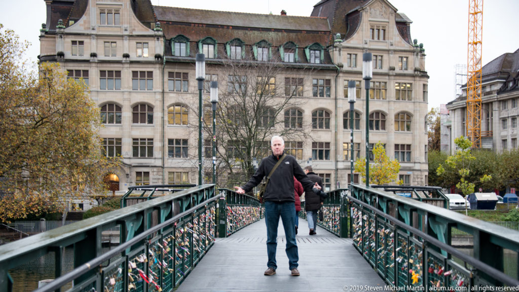 Man walking toward the camera on a metal pedestrian bridge covered with padlocks, with historic Zurich buildings behind.