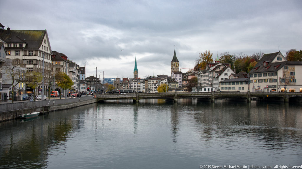 Limmat River through central Zurich with historic buildings on both banks, a bridge, and church towers beneath an overcast sky.