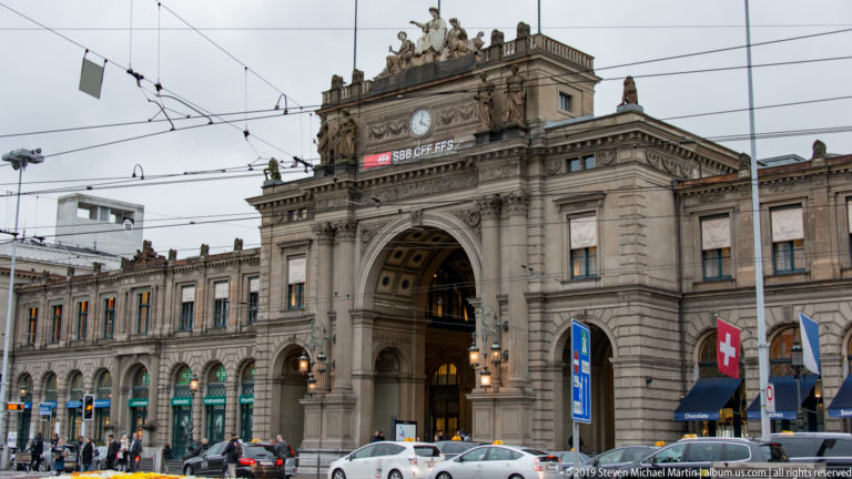 Central arched entrance of Zürich Hauptbahnhof with SBB CFF FFS sign, carved stonework, rooftop sculpture, and tram wires.