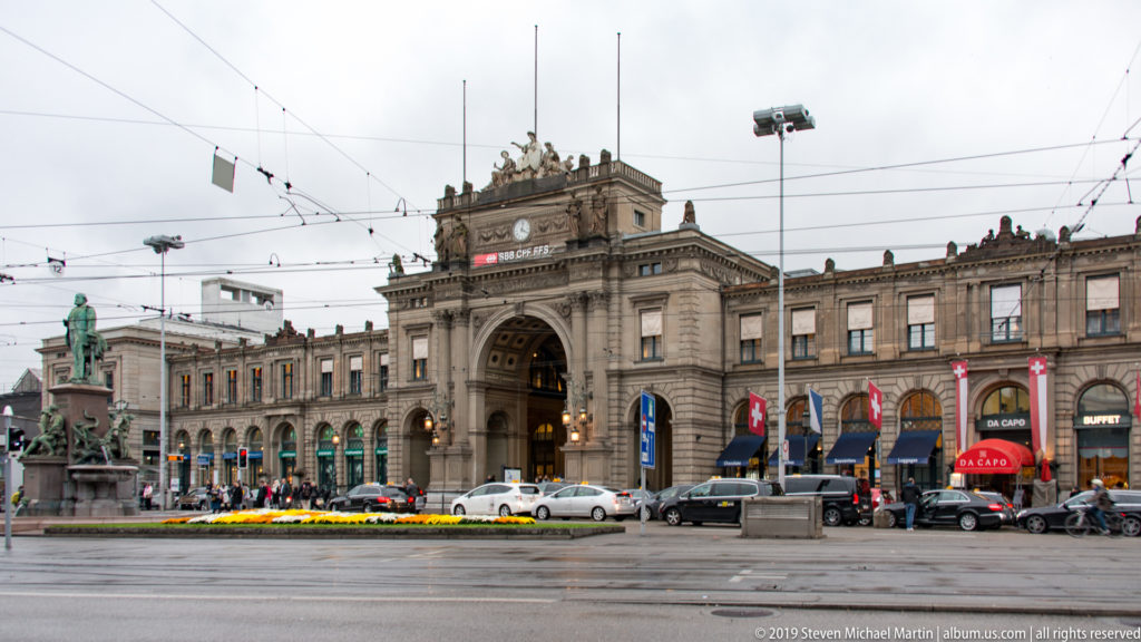 Broad stone façade of Zürich Hauptbahnhof with monumental central arch, rooftop sculpture, Swiss flags, traffic, and tram wires.