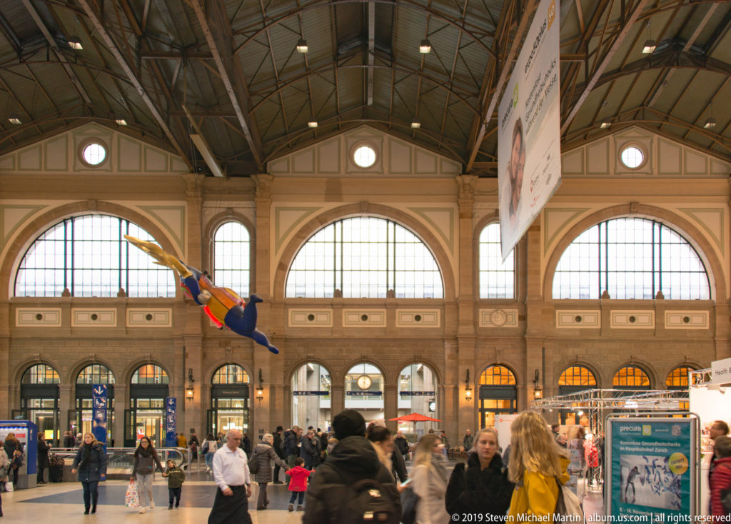 Busy main hall of Zürich Hauptbahnhof with tall arched windows, iron roof structure, shops, travelers, and a suspended colorful figure.
